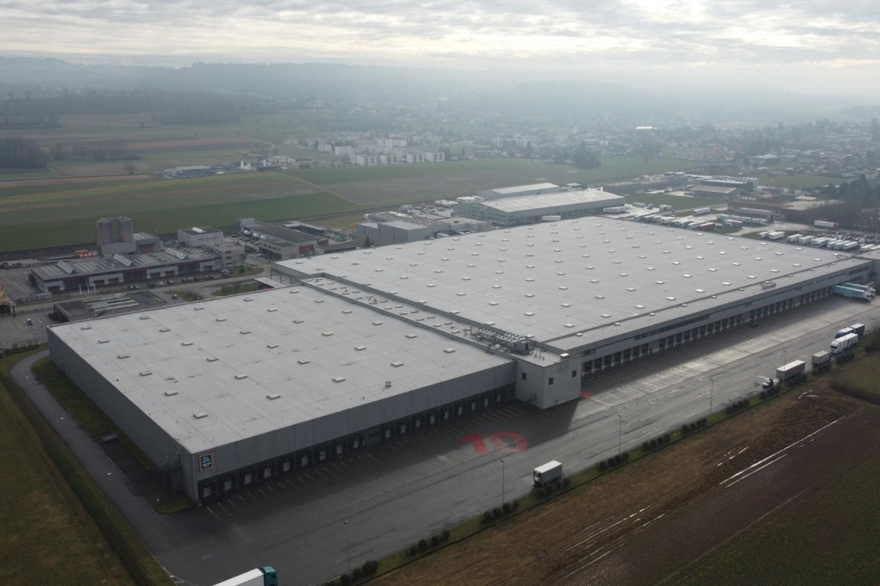 Luftaufnahme eines großen Industriegebäudes mit flachem Dach und mehreren runden Dachaufbauten, umgeben von Feldern und einer Stadt im Hintergrund.
