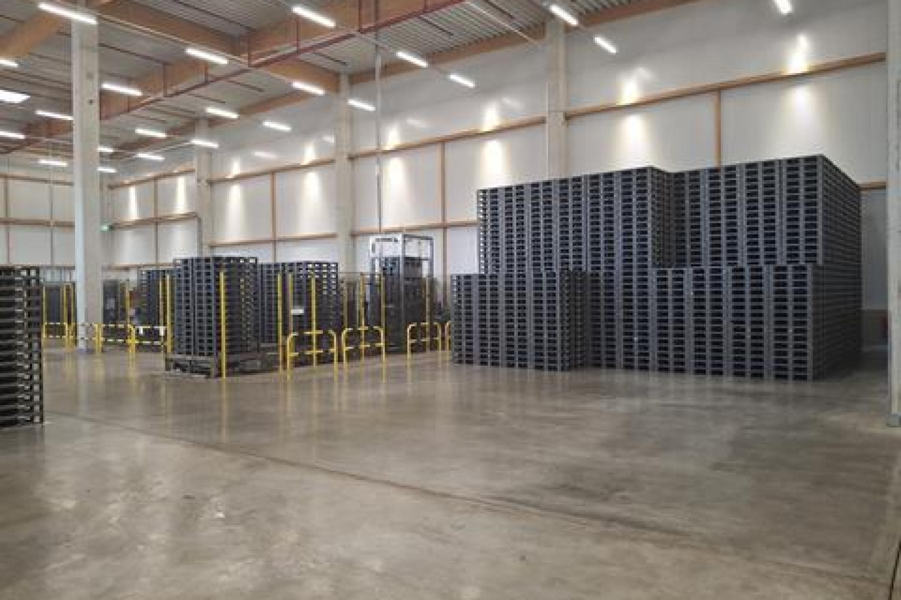Große Lagerhalle mit hohen Decken, mehreren Stapeln schwarzer Kunststoffpaletten und gelben Schutzgeländern.