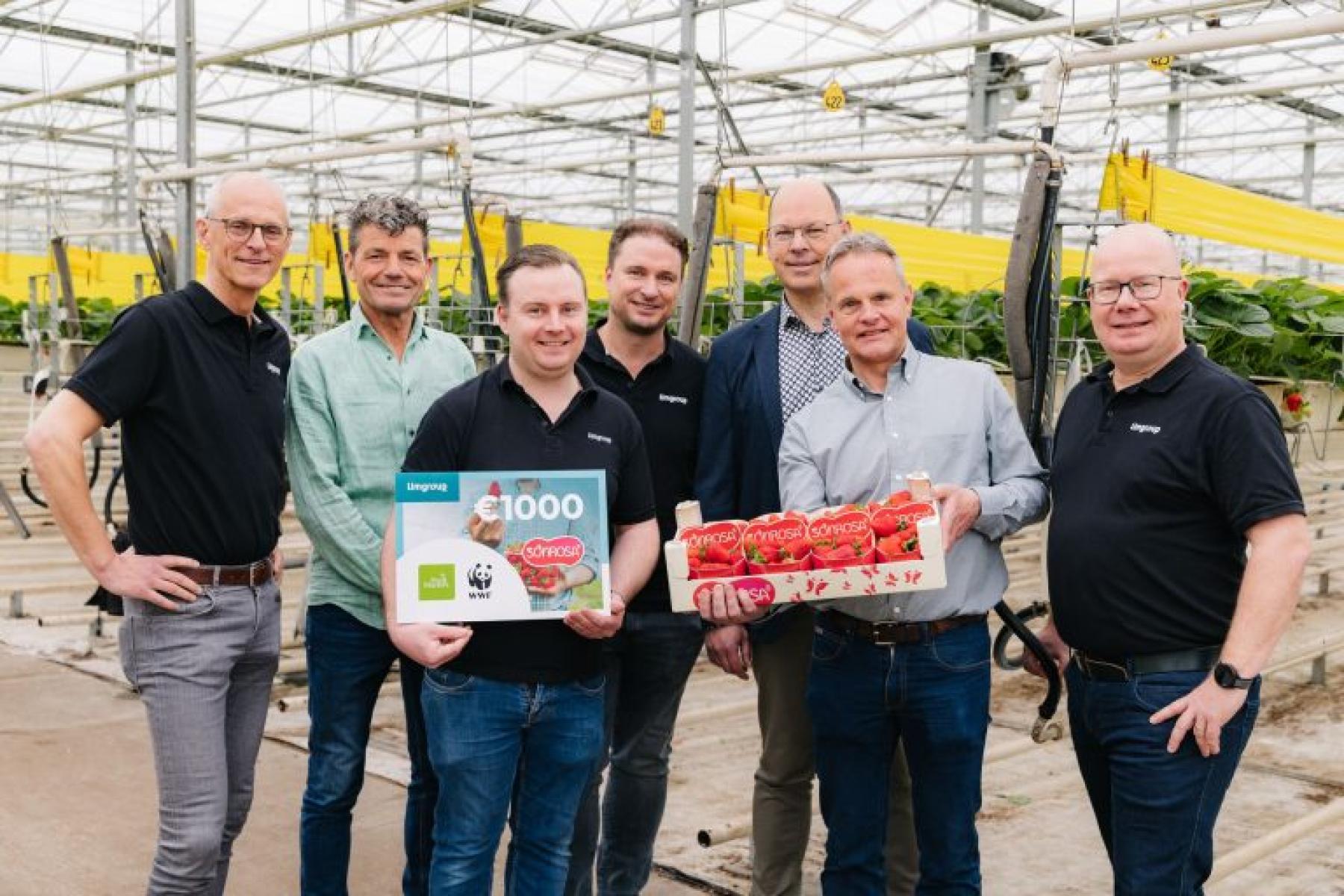 Gruppe von sieben Personen in Gewächshaus, einer hält eine Geschenkkarte und ein weiterer eine Kiste mit Erdbeeren.