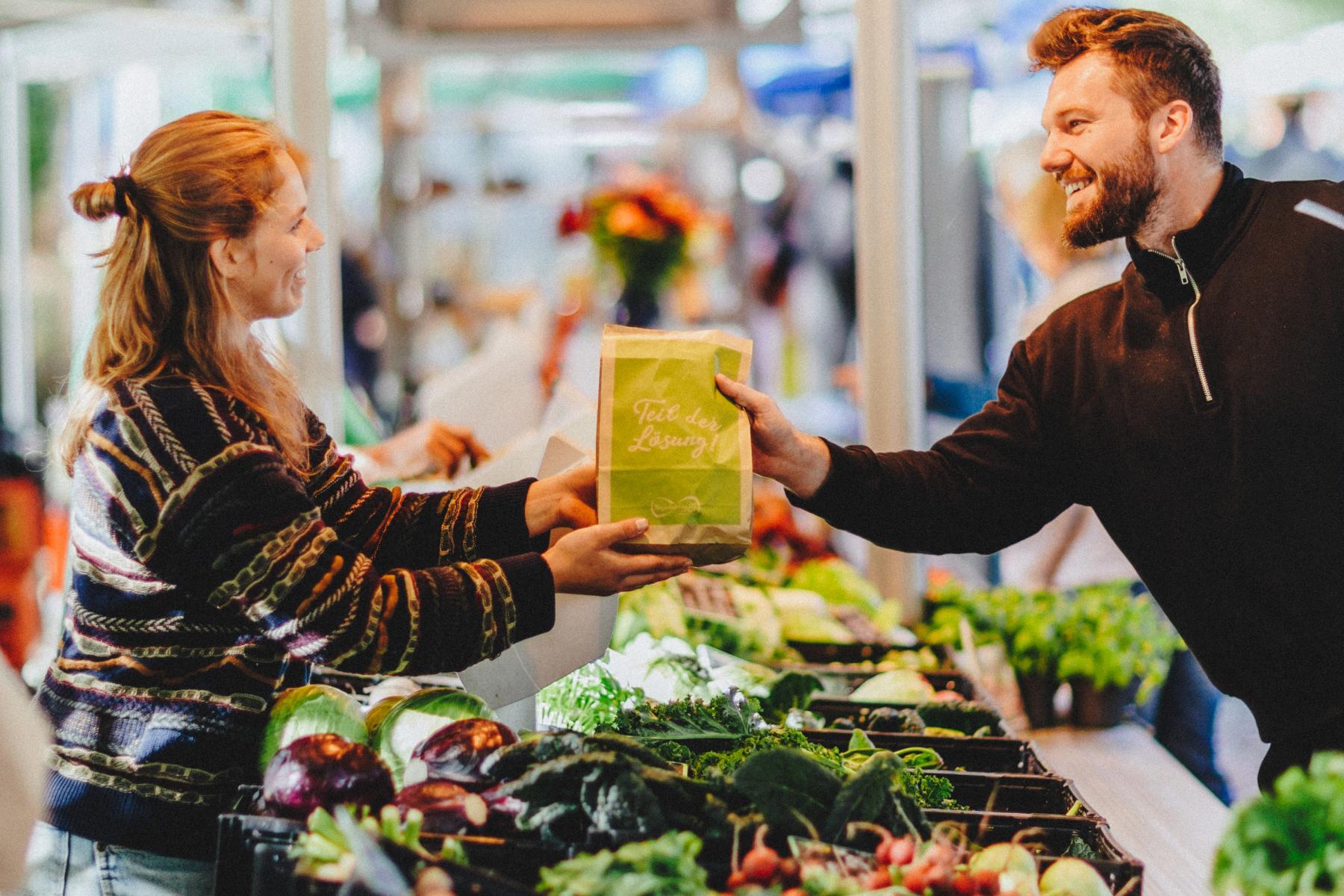 Marktstand an dem ein Mann einer Frau einen Beutel mit Bio-Lebensmittel gibt