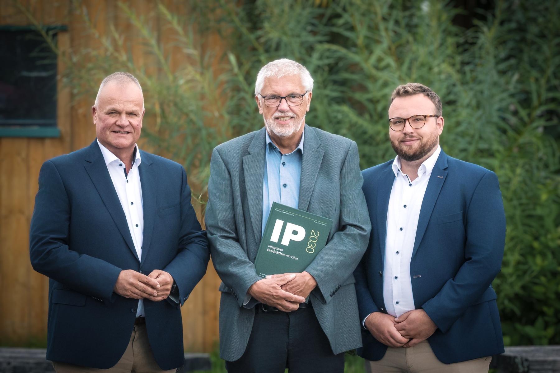 Drei Männer stehen nebeneinander, der in der Mitte hat einen Ordner, auf dem IP steht. 