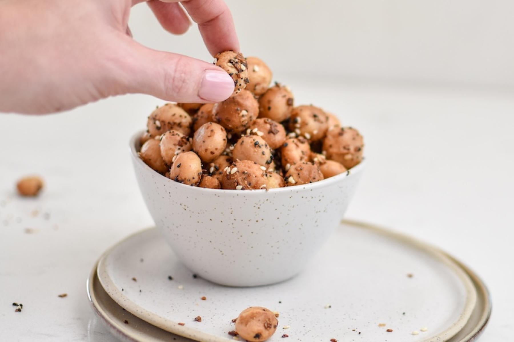 Mit Gewürzen versehene Macadamias in einer weißen Schlüssel. Eine weibliche Hand greift zu. 