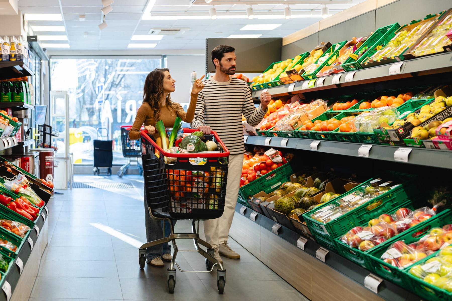 Ein Mann und eine Frau schiebe einen Einkaufswagen durch eine Obst- und Gemüseabteilung.   