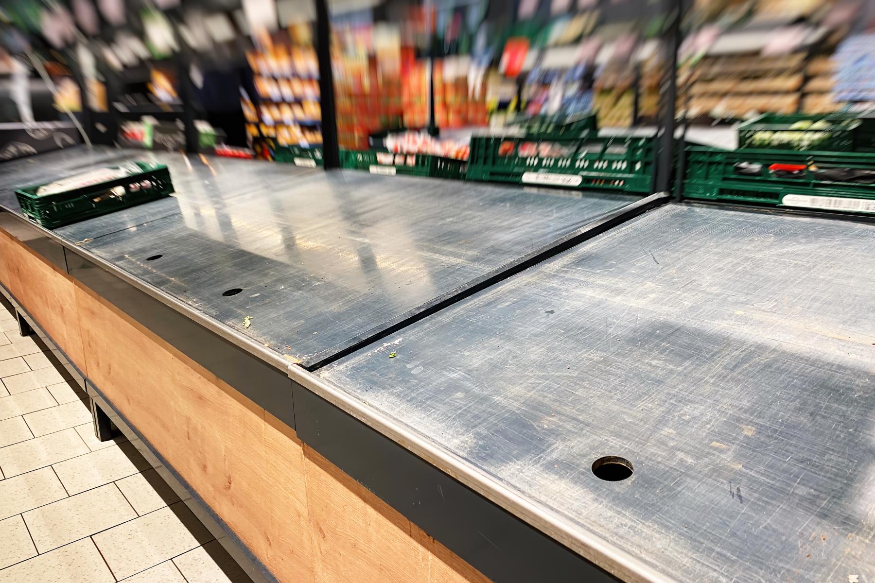 Weitgehend leere Auslage in der Obst- und Gemüseabteilung eines Supermarkts, mit wenigen grünen Warenkisten im Hintergrund.