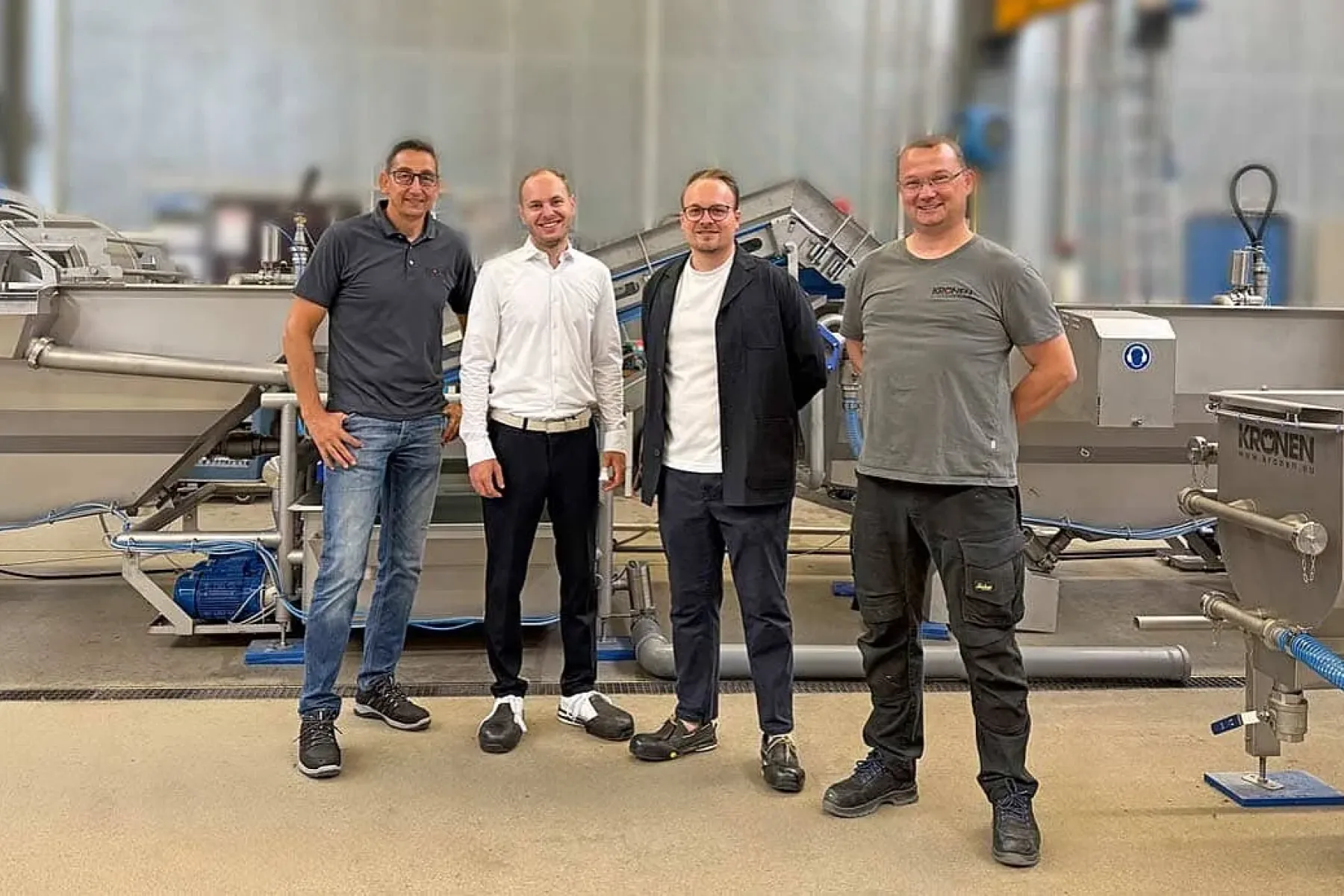 Vier Männer stehen vor einer KRONEN-Salatverarbeitungslinie mit Edelstahlmaschinen und Förderbändern in einer Produktionshalle.