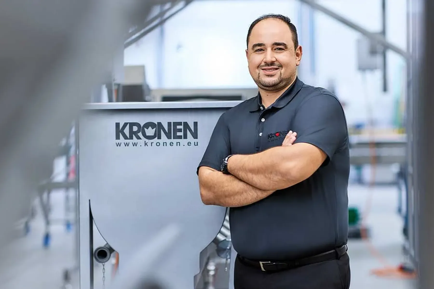 Wissam Badr, Leiter Finanzen und Prokurist der KRONEN GmbH, steht mit verschränkten Armen neben einer Maschine mit KRONEN-Logo.