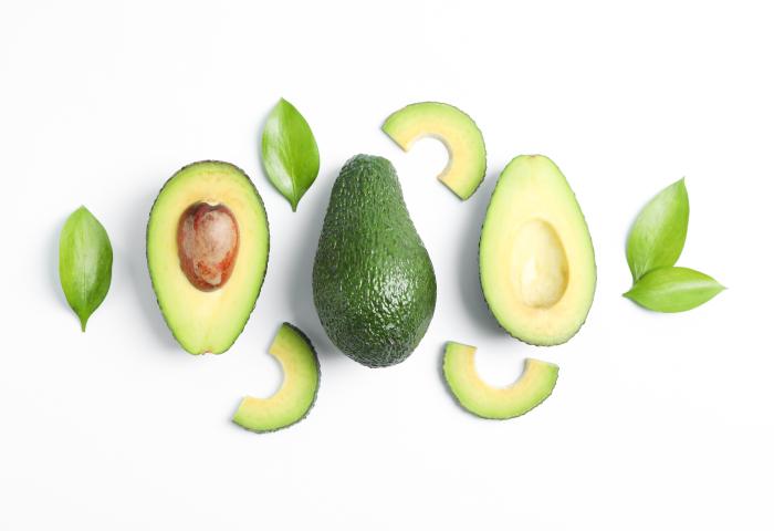 Ganze und halbierte Avocado mit Kern und Scheiben, umgeben von grünen Blättern auf weißem Hintergrund