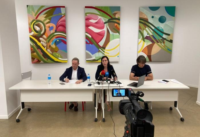 Drei Personen sitzen an einem langen weißen Tisch mit Mikrofonen und Notizen, vor ihnen eine Kamera und hinter ihnen drei abstrakte Gemälde an der Wand