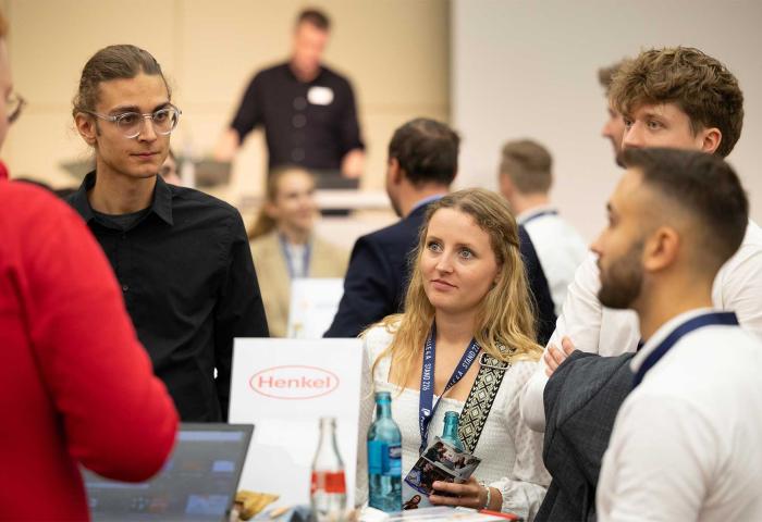 Mehrere Personen stehen und unterhalten sich auf einer Messe, im Vordergrund ein Tisch mit Henkel-Logo und Getränken