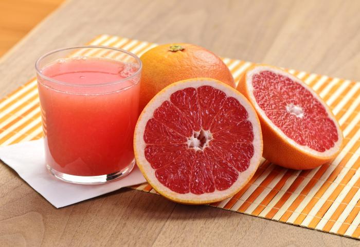 Frisch gepresster Grapefruitsaft im Glas neben einer ganzen und zwei halbierten Grapefruits auf einem gestreiften Untersetzer