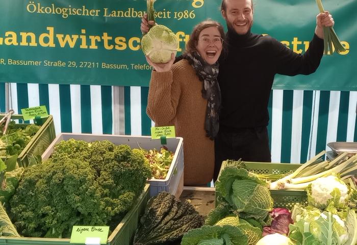 Julia Albrecht und Mitarbeiter Nicolas Hasselbeck stehen hinter einem Marktstand mit frischem Bio-Gemüse wie Grünkohl, Kohlrabi und Lauch, Banner 'Biolandhof Steding Ökologischer Landbau seit 1986' im Hintergrund