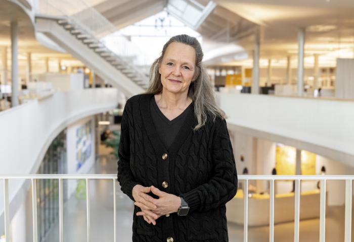 Kirsten Keller steht in einem modernen Bürogebäude mit hellem Innenraum, trägt einen schwarzen Strickcardigan und eine Smartwatch