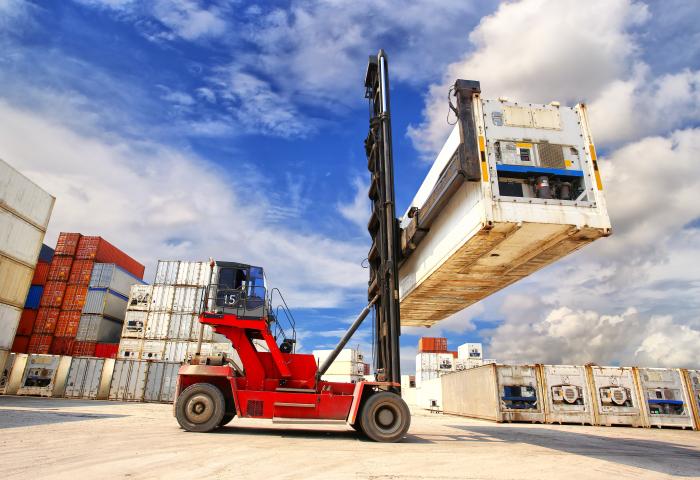 Roter Containerstapler hebt einen weißen Frachtcontainer in einem Hafen mit gestapelten Containern und blauem Himmel