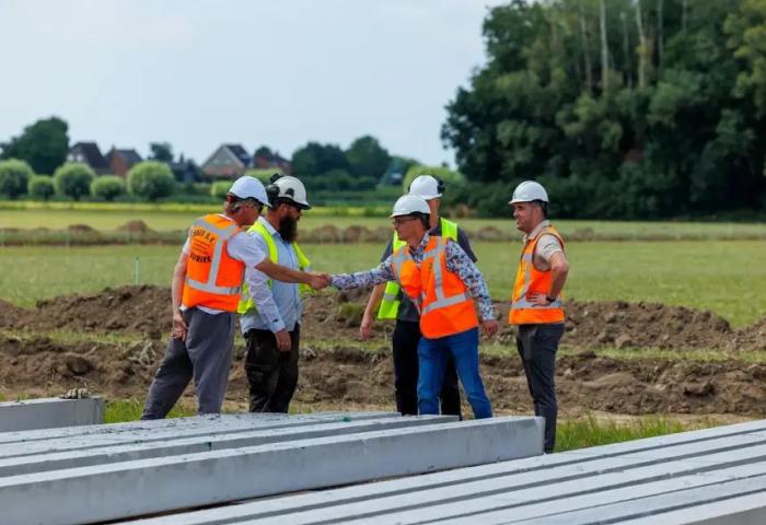 Fünf Personen auf der Baustelle. Sie tragen Schutzhelme und orange Warnwesten, zwei schütteln sich die Hand