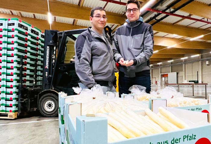 Hugo da Motto und Leo Zastrow vom Spargelbüro der Pfalzmarkt eG stehen in einer Lagerhalle neben Kisten mit frischem Spargel aus der Pfalz