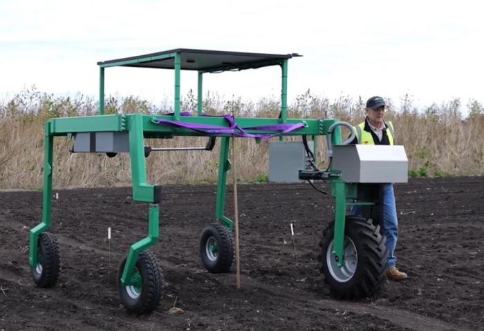 Prof. Daniel Rodriguez steht neben dem RootBot, einem großen grünen landwirtschaftlichen Roboter mit vier Rädern auf einem Acker