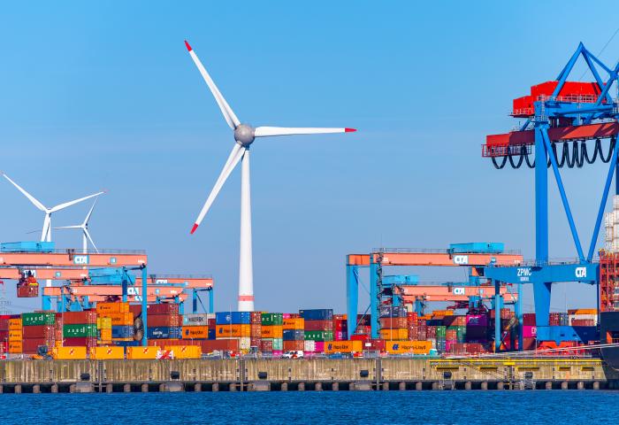 Containerterminal mit mehreren bunten Containern, einem großen Kran und einem Windrad im Hintergrund. Wasser im Vordergrund.