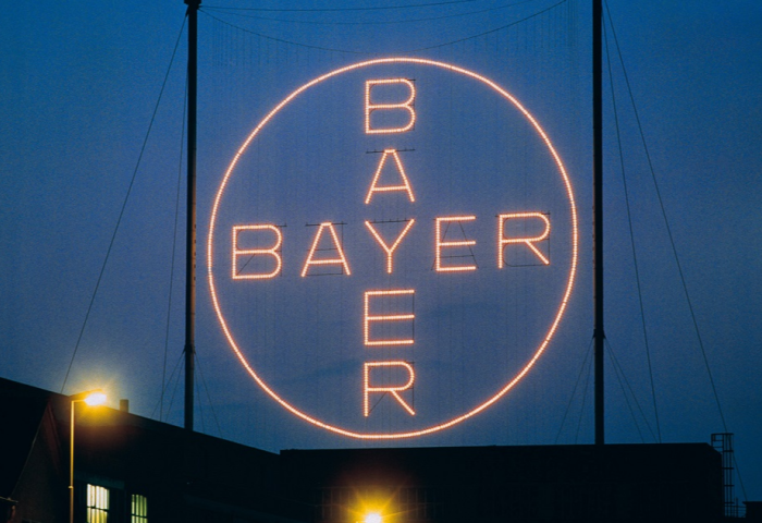 Leuchtendes Bayer-Kreuz mit dem Schriftzug 'BAYER' in einem Kreis, nachts vor dunklem Himmel