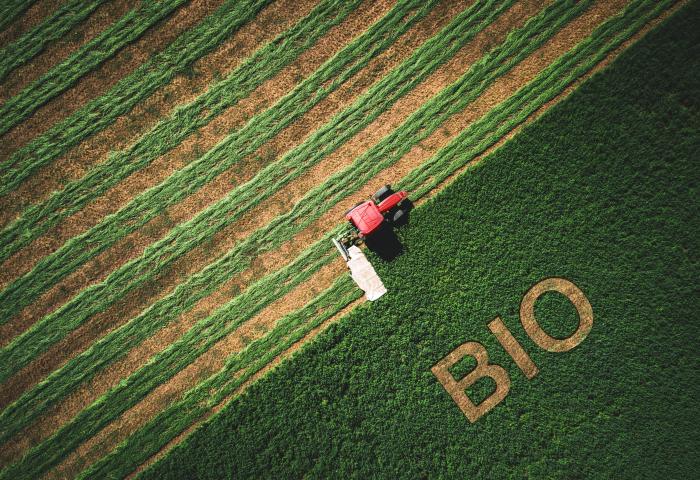Luftaufnahme eines roten Traktors auf einem Feld mit grünen und braunen Streifen, daneben ist das Wort 'BIO' in das Gras geschnitten.