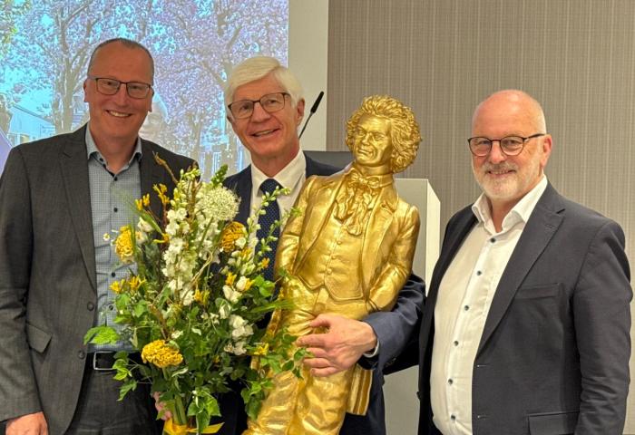 Jürgen Bruch, Dieter Krauß und Dr. Andreas Brügger stehen zusammen, halten eine goldene Statue und einen Blumenstrauß. Im Hintergrund ist eine Präsentation sichtbar.