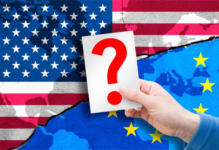 Hand hält Karte mit rotem Fragezeichen vor zerrissener US-Flagge und EU-Flagge, symbolisiert Ungewissheit in Beziehungen