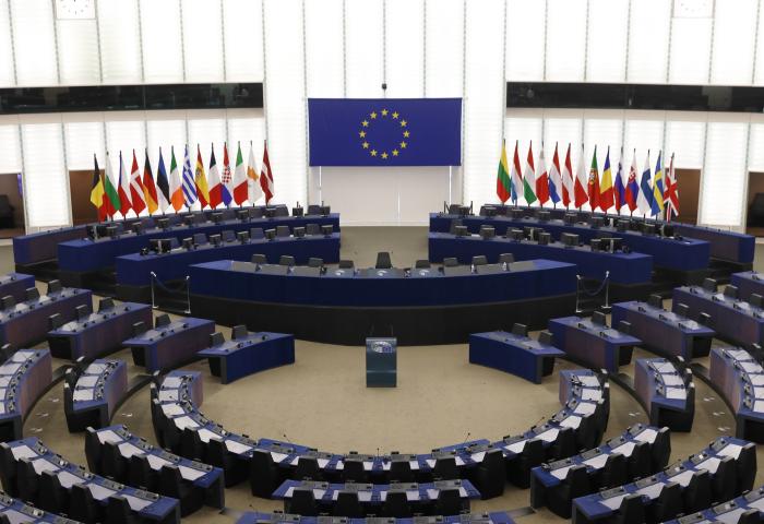 Leerer Plenarsaal des Europa Parlaments mit halbkreisförmigen Sitzreihen, Rednerpult und EU-Flagge an der Wand, flankiert von mehreren Nationalflaggen