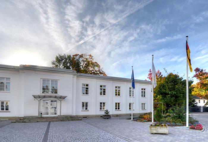 Gebäude des Bundeskartellamtes, weißes Verwaltungsgebäude mit mehreren Fenstern, drei Flaggenmasten mit EU-, Deutschland- und Bundeskartellamtsflagge vor dem Eingang