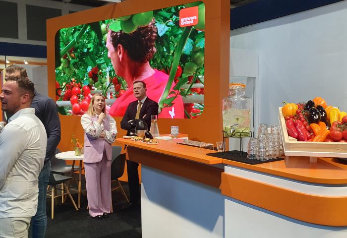 Messestand von Growers United mit großem Bildschirm, der eine Frau vor Tomaten zeigt, und einem Tresen mit frischem Gemüse und Getränkespender