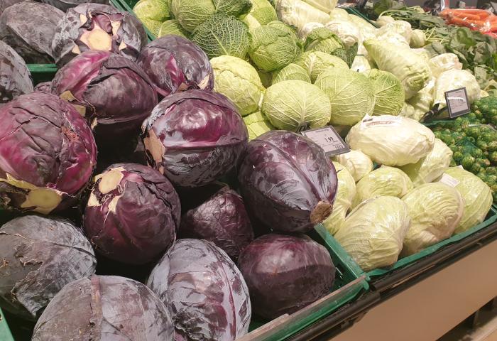Mehrere Köpfe Rotkohl und Weißkohl liegen in grünen Kisten im Supermarkt, im Hintergrund Karotten und Rosenkohl sichtbar.