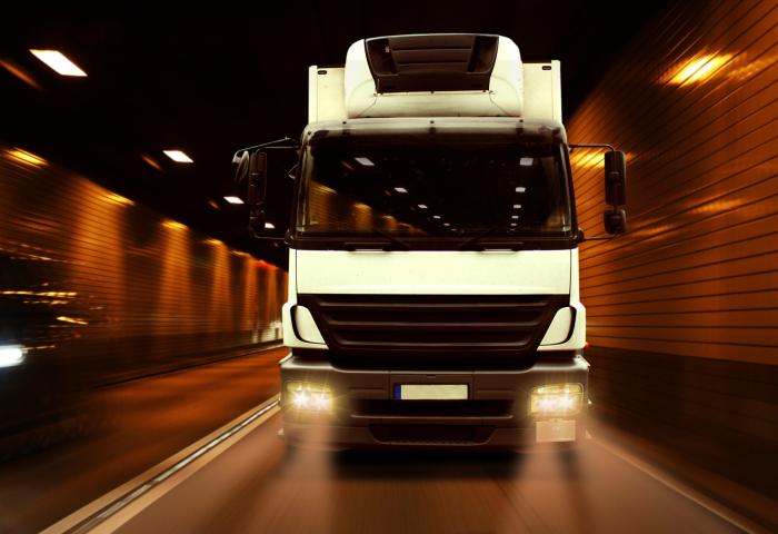 Weißer LKW mit eingeschalteten Scheinwerfern fährt frontal durch einen beleuchteten Tunnel