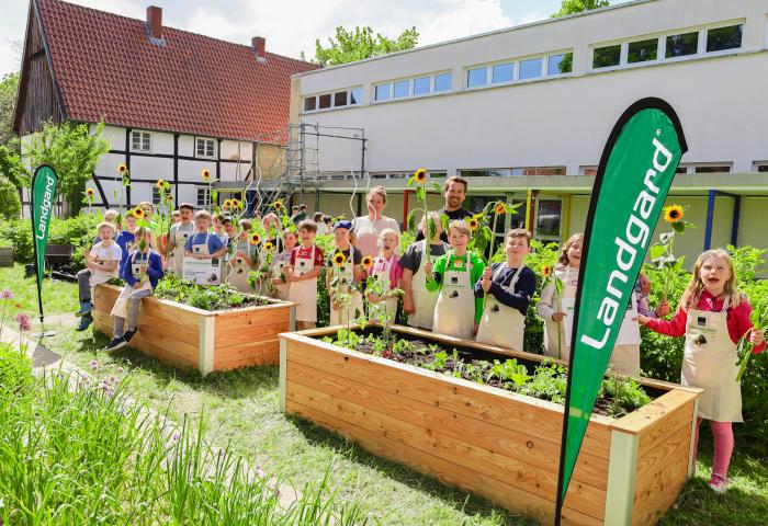 Gruppe von Kindern in Schürzen, die in einem Schulgarten stehen, umgeben von Hochbeeten mit Sonnenblumen und Gemüse. Zwei grüne Fahnen mit 'Landgard'
