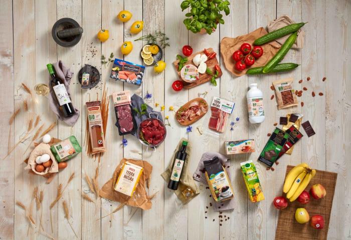 Verschiedene Lebensmittel auf hellem Holztisch, darunter Wein, Eier, Bio-Fleisch, Gemüse, Obst, Nudeln, Milchprodukte und Säfte, arrangiert in Gruppen.