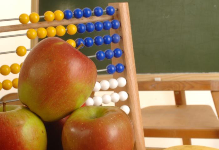 Drei rote Äpfel vor einem Abakus mit gelben, blauen und weißen Perlen in einem Klassenzimmer