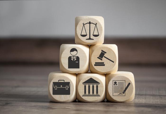 Fünf hölzerne Würfel mit schwarzen Symbolen: Waage, Richter, Hammer, Aktentasche, Gerichtsgebäude und unterschriebener Vertrag, gestapelt auf Holztisch.