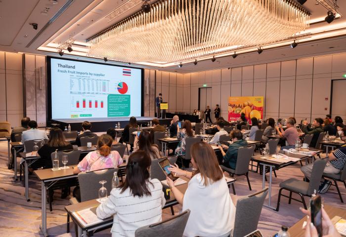 Konferenzraum mit Teilnehmern, die einer Präsentation über Frischobstimporte in Thailand folgen, Sprecher auf dem Podium vor großem Bildschirm