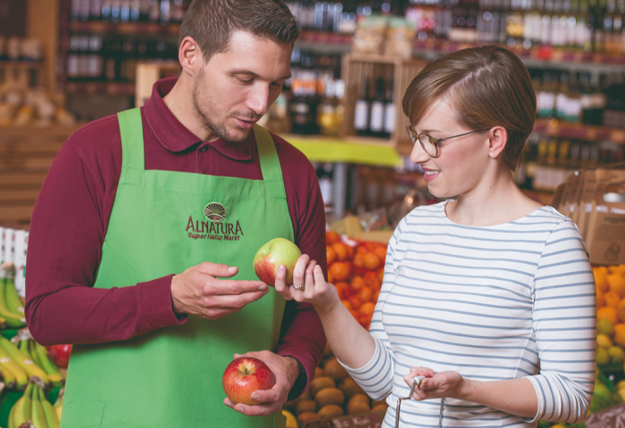 Mitarbeiter im Alnatura Super Natur Markt zeigt Kundin zwei Äpfel im Obst- und Gemüseregal