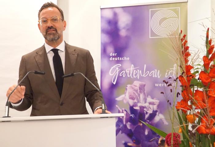 Bertram Fleischer steht hinter einem Rednerpult, neben einem Banner mit lila Blumen und Text 'der deutsche Gartenbau ist wert'