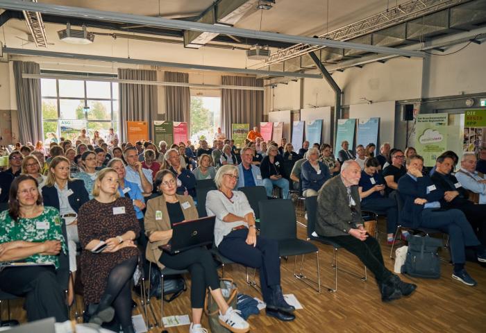Große Gruppe von Menschen sitzt in einem hellen Konferenzraum und hört einem Vortrag zu, viele mit Namensschildern und Notizmaterialien