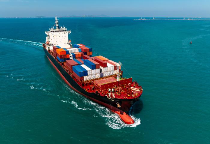 Containerschiff mit bunten Frachtcontainern fährt auf offenem Meer unter klarem Himmel, Küstenlinie im Hintergrund sichtbar