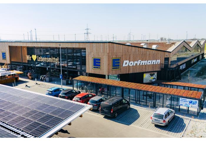 Edeka Dorfmann Supermarkt mit Holzfassade und großen Fenstern, mehrere Autos auf dem Parkplatz, sichtbare Solarpanels auf dem Dach.