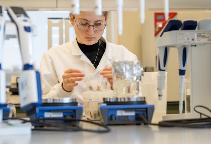 Person in weißem Laborkittel sitzt an einem Labortisch mit Laborgeräten und hält ein kleines Objekt in der Hand.