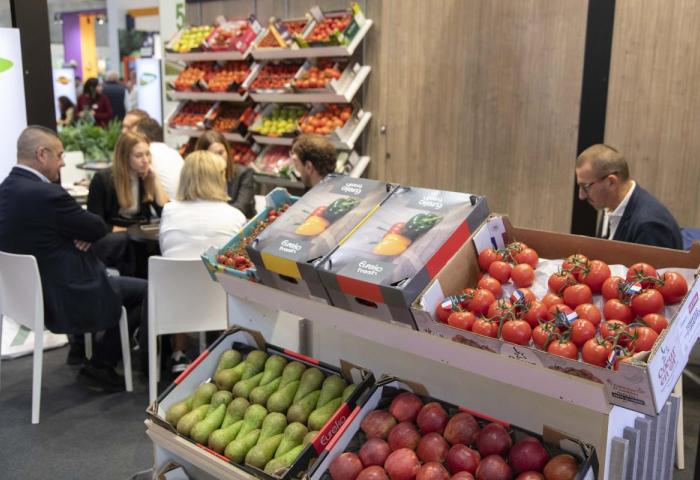 Mehrere Personen sitzen an einem Tisch in einem Messebereich, im Vordergrund Kisten mit Tomaten, Avocados und Granatäpfeln