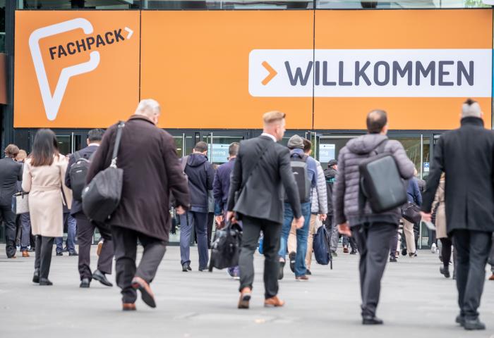 Menschen gehen auf den Eingang der Fachpack-Messe zu, großes orangefarbenes Schild mit 'Fachpack' und 'Willkommen'