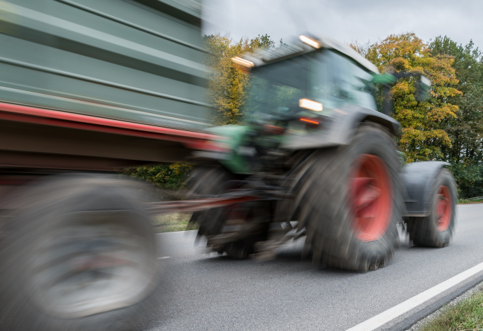 Fahrender Traktor mit Anhänger auf Landstraße, unscharf durch Bewegung, Bäume im Hintergrund