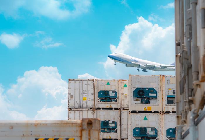 Frachtflugzeug mit der Aufschrift 'CARGO' hebt über gestapelten weißen Kühlcontainern mit blauen Kühlaggregaten ab, blauer Himmel mit Wolken