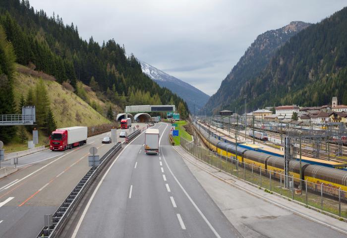Autobahn mit Lastwagen und Autos am Brennerpass zwischen bewaldeten Bergen und einem Bahnhof mit Güterzügen