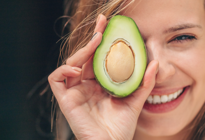 lächelnde Frau hält sich eine halbierte Avocado mit sichtbarem Kern vors Auge