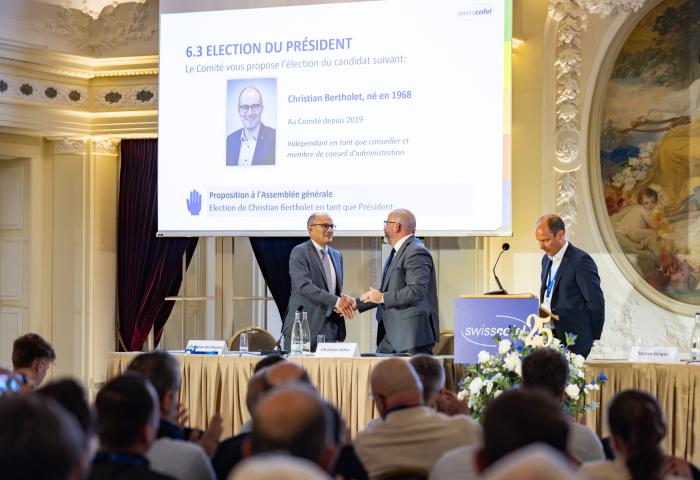 Christian Sohm gratuliert Christian Bertholet zur Wahl als Präsident, beide stehen, im Hintergrund ein Bildschirm mit Wahlinformationen.