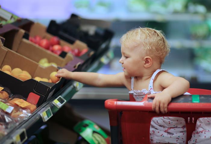 Kleinkind sitzt in einem Einkaufswagen und greift in die Obstauslage eines Supermarktes