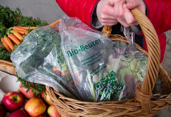 Hände halten Korb mit kompostierbaren Bio-Beuteln aus nachwachsenden Rohstoffen, umgeben von frischem Obst und Gemüse